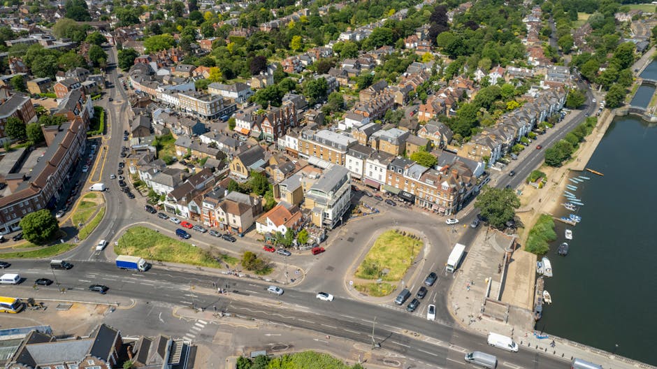 An aerial view of a town showing a busy street intersection adjacent to a river, with several parked and moving vehicles including trucks and vans. The image features a mix of residential buildings, c